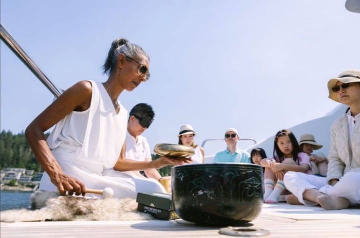 Passengers sit on a boat deck in sunlight as a singing bowl ceremony takes place beside calm shoreline water.