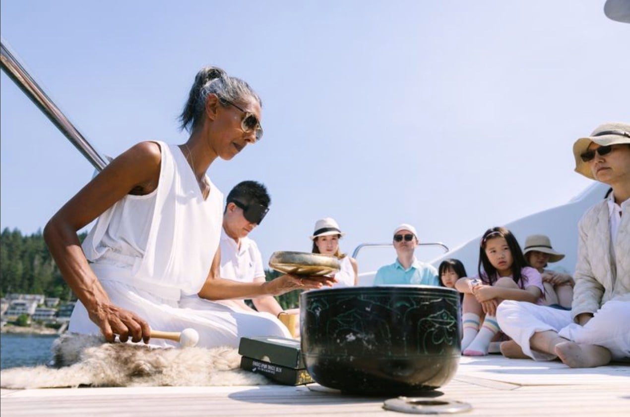 Passengers sit on a boat deck in sunlight as a singing bowl ceremony takes place beside calm shoreline water.