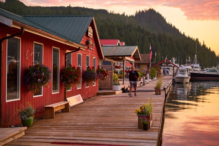 Wooden dock lined with red buildings and flower baskets, boats moored at sunset against forested hills.