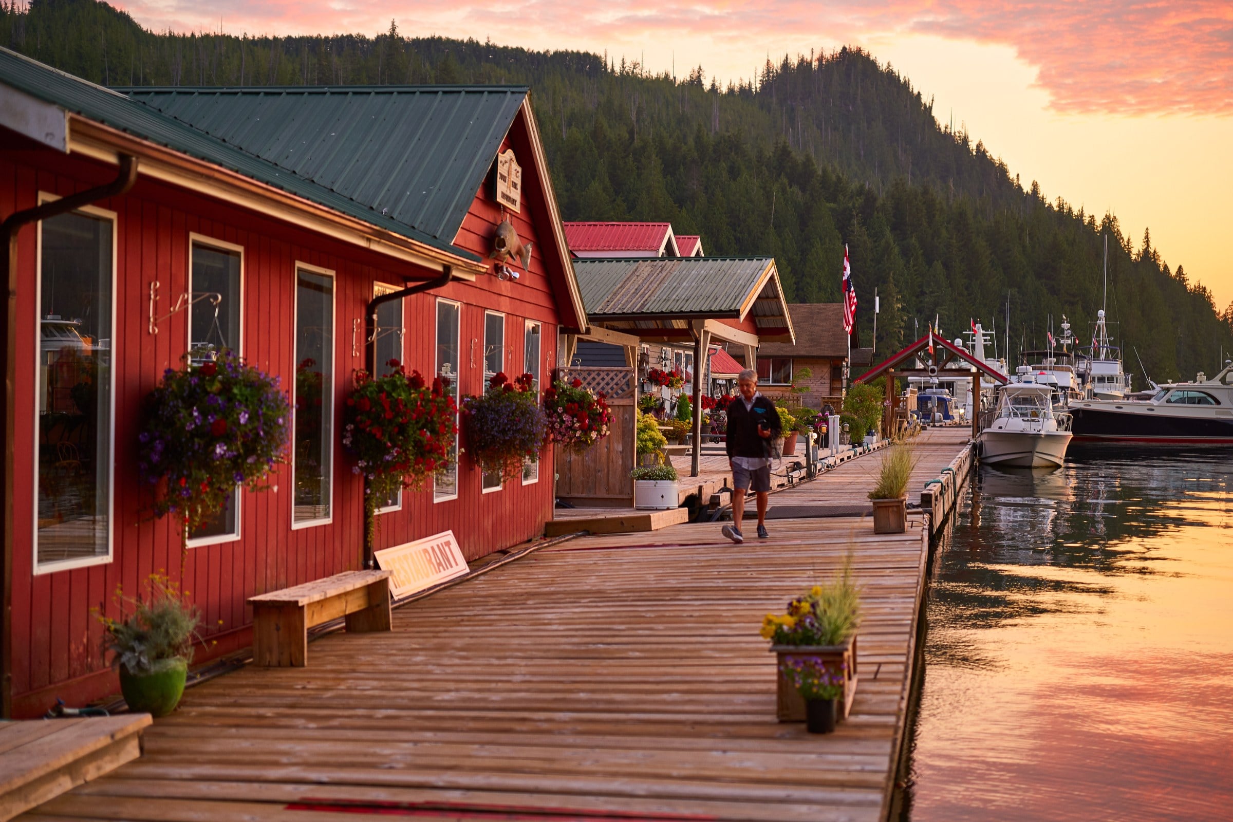Wooden dock lined with red buildings and flower baskets, boats moored at sunset against forested hills.