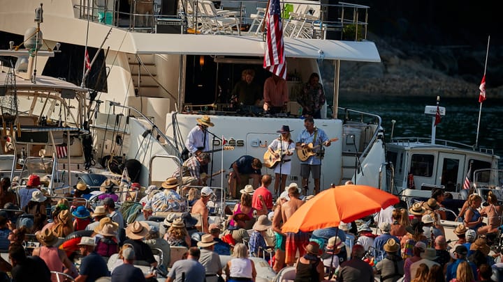 Musicians perform from a yacht's swim platform to a crowd of boaters gathered under hats and umbrellas on the water.