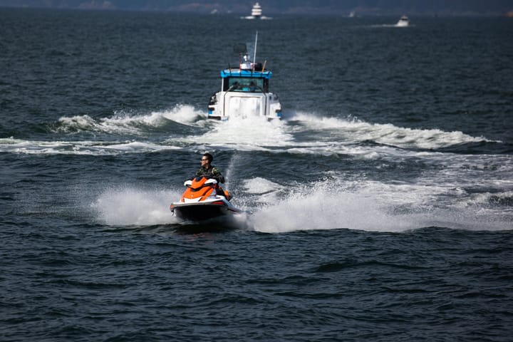 A jet ski cuts across choppy water ahead of a white boat throwing up spray under open sky.