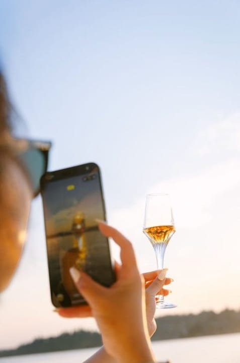 A phone photographs a glass of golden wine against soft evening light over calm water.