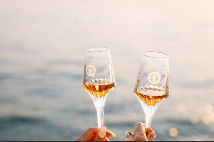 Two champagne glasses clink together above calm open water catching soft evening light.