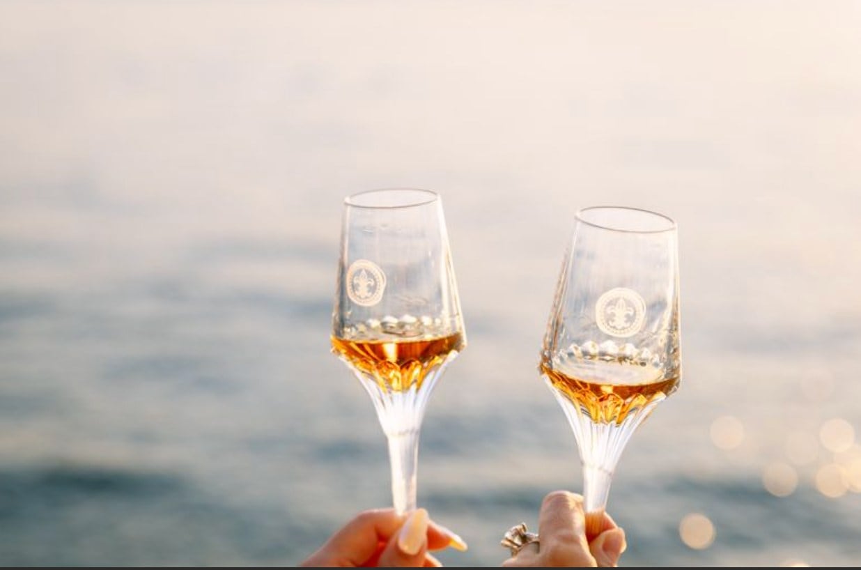 Two champagne glasses clink together above calm open water catching soft evening light.