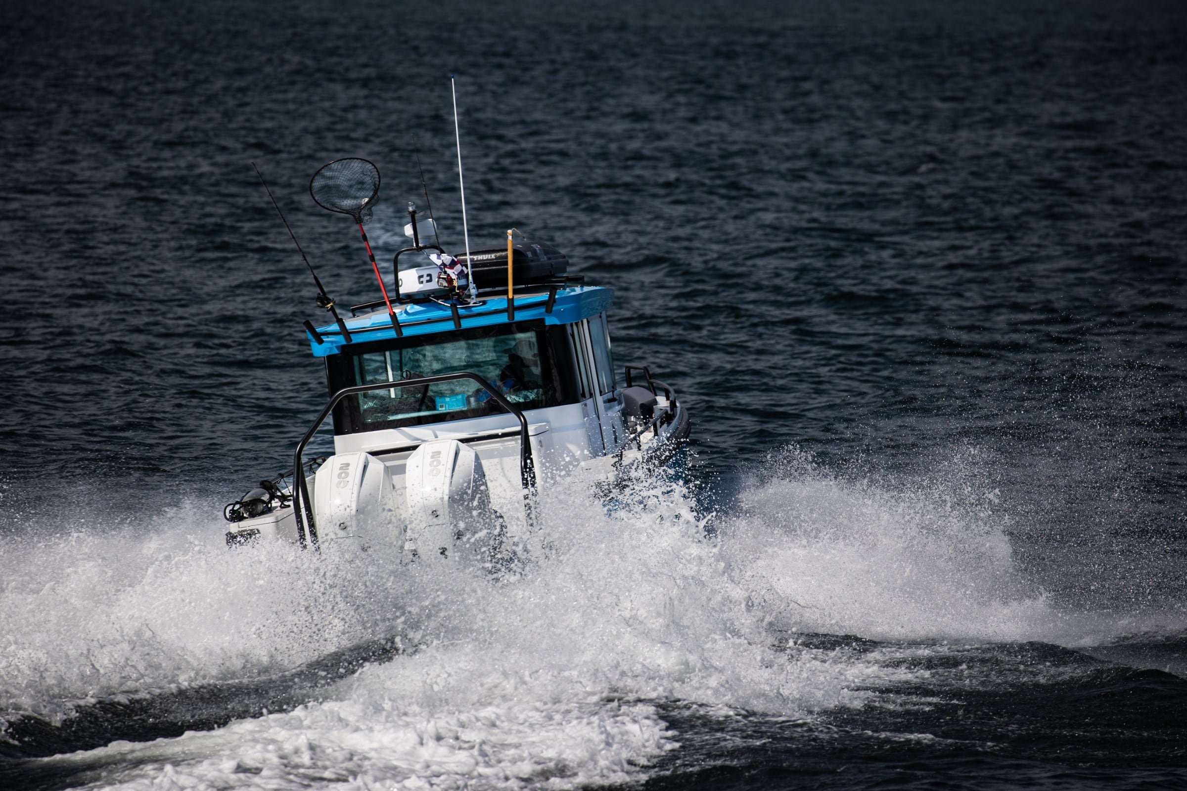 A white and blue cabin boat with triple outboards cuts through dark water, throwing spray on both sides.