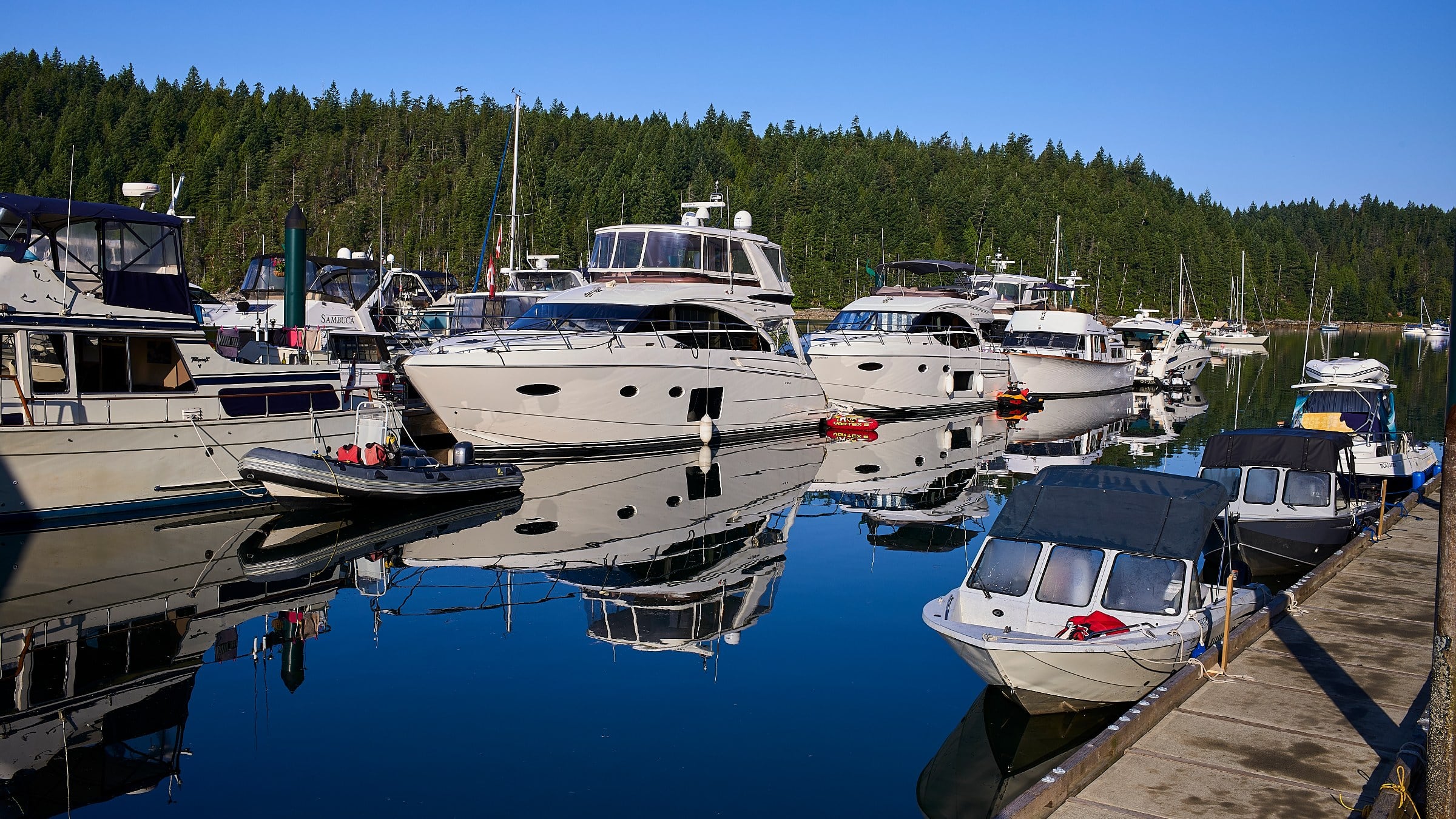 Yachts and small boats moored at a dock, reflected in still water beneath forested hills.