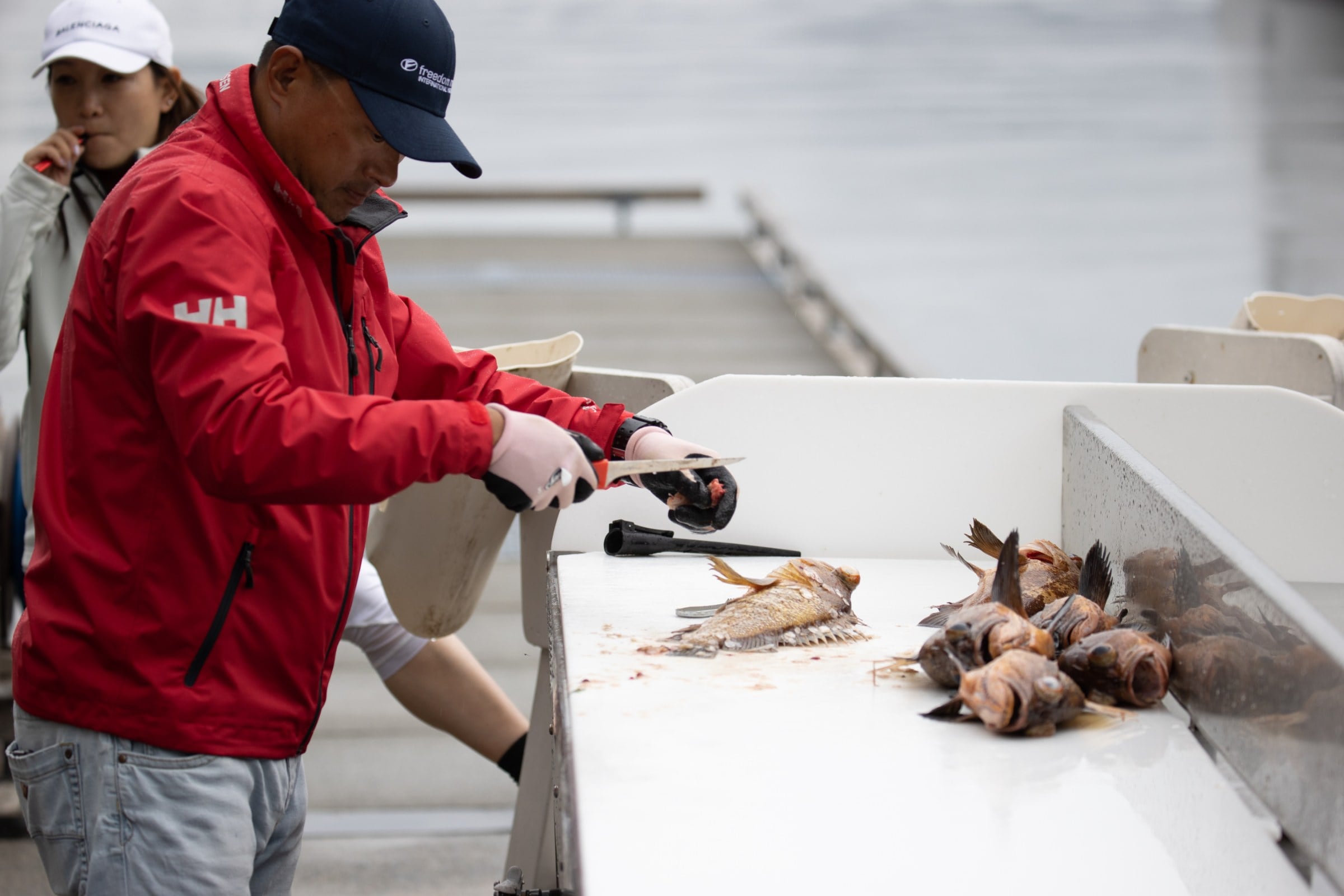 Someone in a red jacket cleans freshly caught fish on a dockside table beside calm grey water.